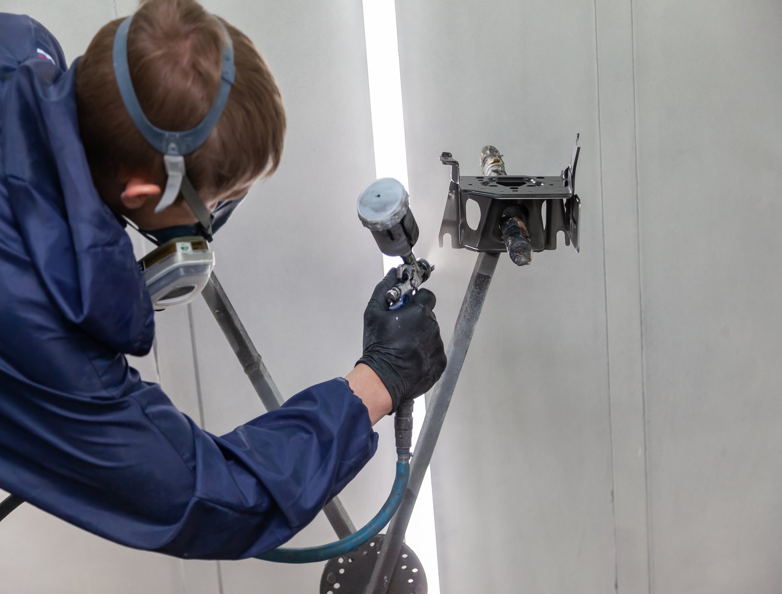 A male worker in jumpsuit and gloves paints with a spray gun a part of the car body frame in black after being damaged at an accident. Auto service industry professions