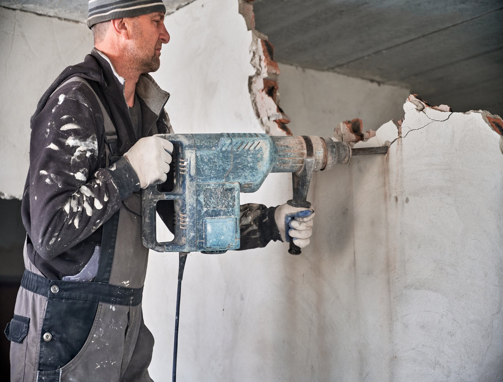 Man builder in workwear drilling wall with electric hammer drill. Male worker using electric drill while breaking wall in apartment under renovation. Demolishing and construction destroying concept.