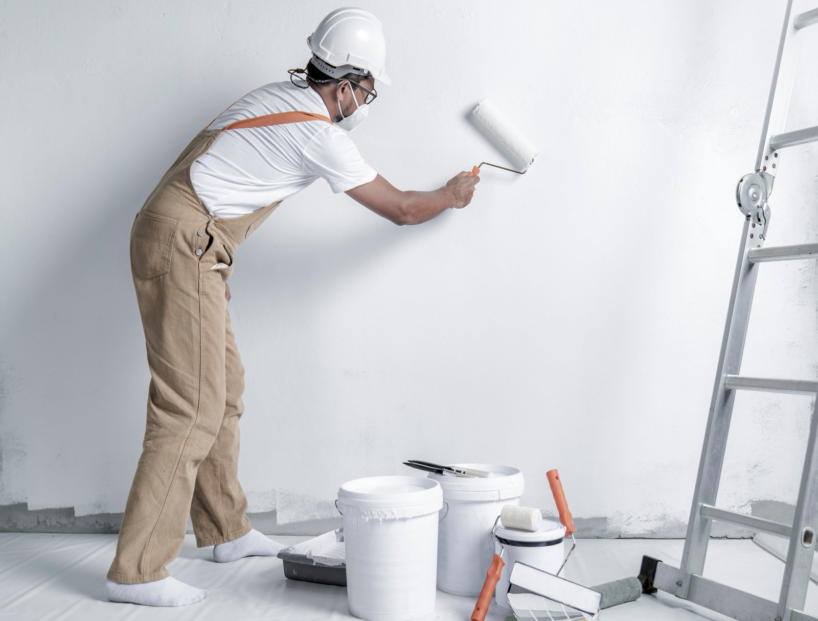 a man paints a white wall with a roller. Repair of the interior. Young male decorator painting a wall in the empty room, concept builder or painter in helmet with paint roller over the empty room.